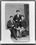 [Unidentified group of three men, full-length portrait, two seated, one standing in the background, facing front]